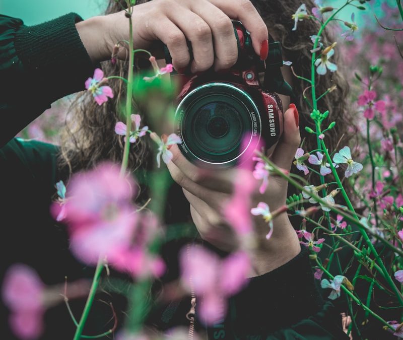 selective focus of woman behind pink flowers holding red Samsung bridge camera about to take photo of flower